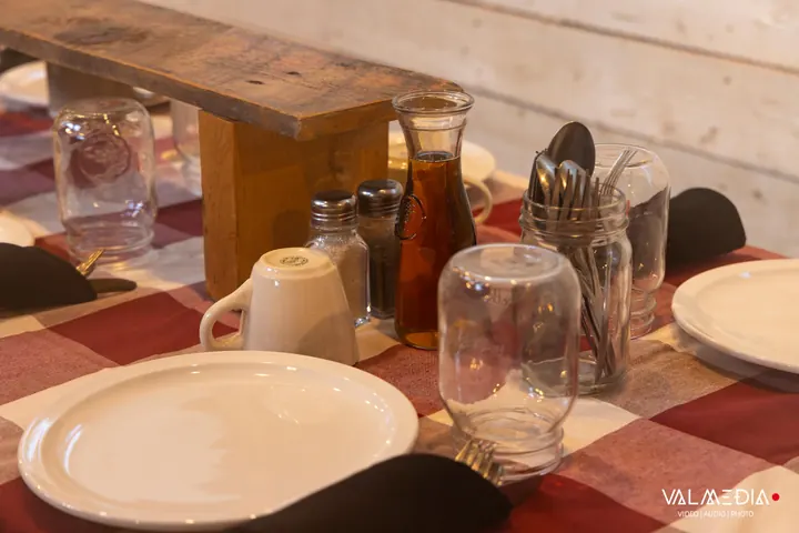 Une table dressée : carafe de sirop d'érable, pots de verre avec les ustensiles et assiettes blanches sur une nappe à carreaux.