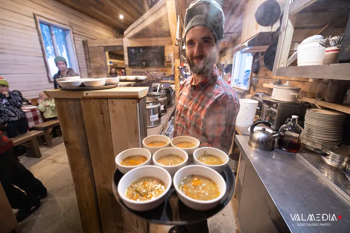 A smiling server carries a tray of six steaming bowls of soup through the shack's dining room
